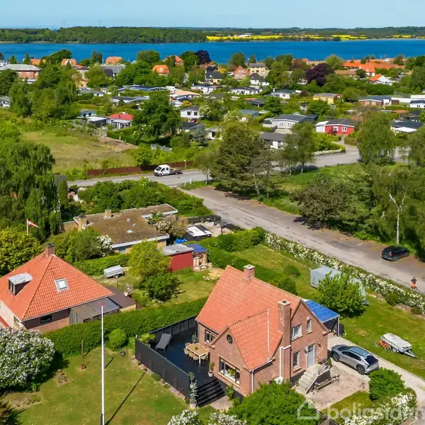 Hus med rød teglstens tag står i en frodig have. Det er omgivet af grønne områder og andre huse. I baggrunden ses en sø eller fjord med træer på bredden.