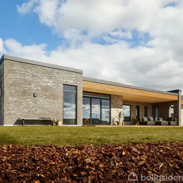 Hus med fladt tag og store vinduer, stående på en græsplæne med barkflis i forgrunden, omgivet af en åben, skyfyldt himmel.