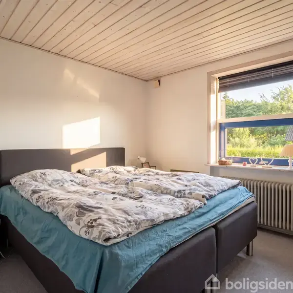 Seng står i et lyst soveværelse med hvidt træloft. Vinduet til højre viser udsigt til have med grønt løv. Natbord med lampe står ved siden af sengen.