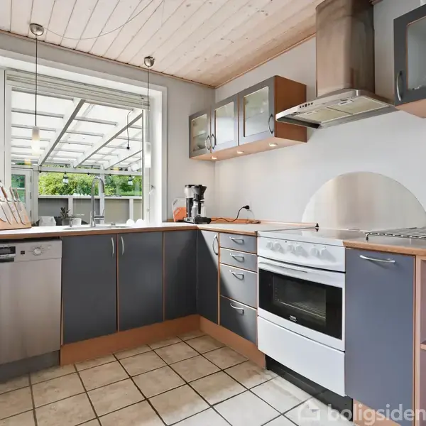 En moderne køkken med grå skabe og hvidevarer. Komfuret er placeret under en emhætte. Bordpladerne er træfarvede. Et stort vindue giver udsigt til en udendørs terrasse. Skabe indeholder køkkenredskaber.