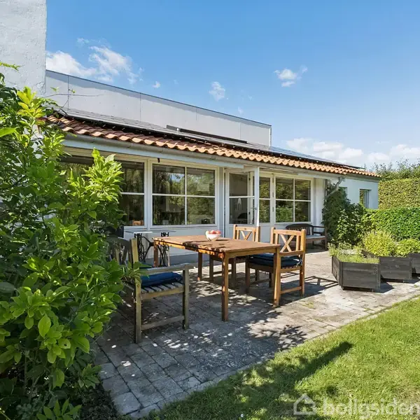 Stue med sort sofa, pejs der brænder. Store vinduer giver udsigt til grønt haveområde. Skrivebord med plante til venstre, reol fyldt med bøger i baggrunden. Ingen tekst.