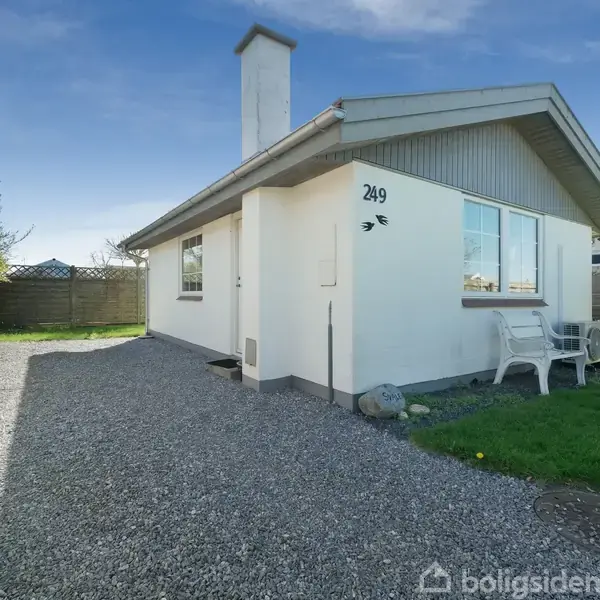 Et hvidt hus står på en grusbelagt sti omgivet af en grøn græsplæne. Nummeret "249" er synligt på huset, som har hvide plaststole ved siden.