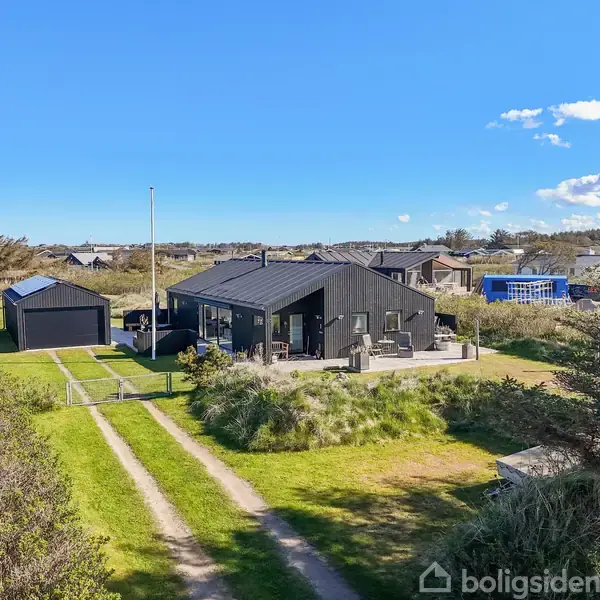 Et sort sommerhus med terrasse står i et grønt, åbent landskab med blå himmel i baggrunden. En grusvej fører op til huset. Rundt omkring ses andre huse i det fjerne.