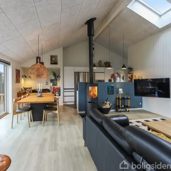 Stue med sort lædersofa, der vender mod en vægmonteret fladskærm. Ved siden af er et spisebord af træ under en stor lampe. Rummet har ovenlys og store vinduer.