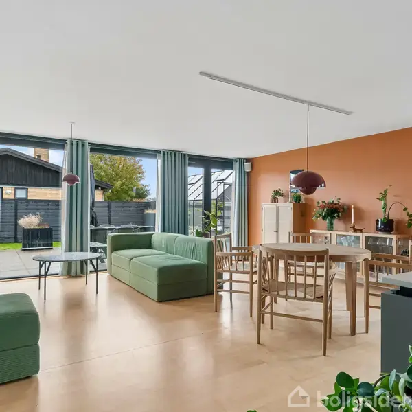 Stue med grønne sofaer og træspisebord, indrettet i moderne stil. Store vinduer med udsigt til have og skur. Lyse gardiner og orange væg som accent.