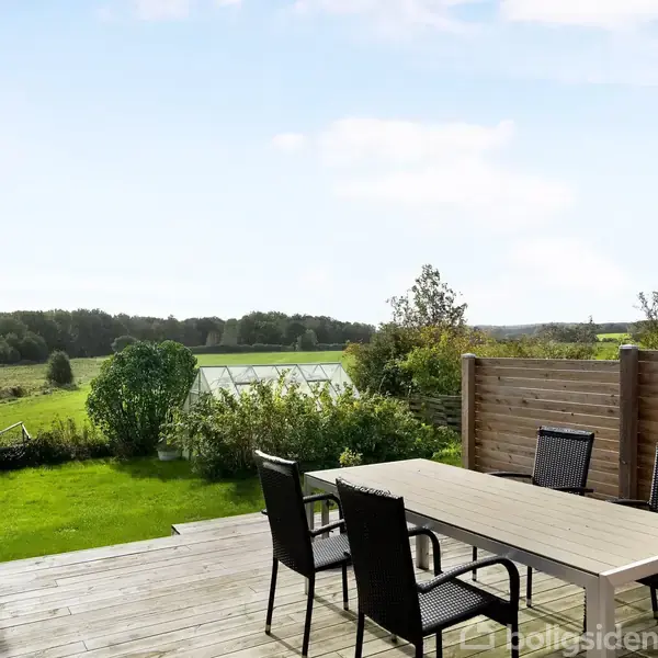 Et bord med fire stole står på en terrasse. I baggrunden ses en have med græs, buske og et drivhus, omgivet af frodig natur under en klar himmel.