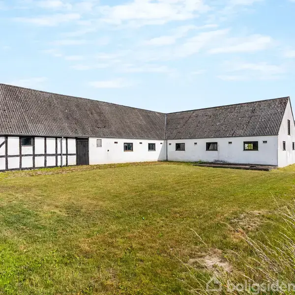 Hvid bindingsværkbygning står stille på en grøn græsplæne, omgivet af landskab med træer og marker under en blå himmel.