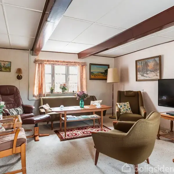 Stue med sofa og lænestole omkring et lavt sofabord. Fjernsyn i hjørnet, billeder på væggene, en lampe ved siden af sofaen, og blomsterdekorationer på bordet.