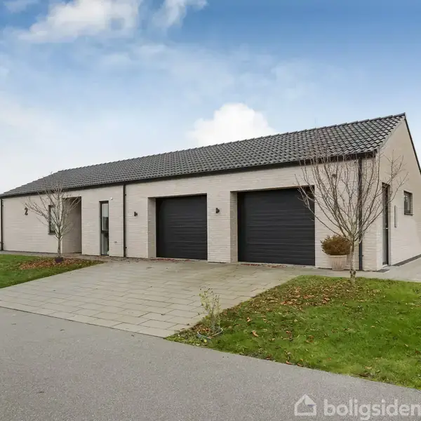 En moderne, lav bygning med to sorte garageporte og indgangsdør. Bygningen står på en flisebelagt indkørsel omgivet af en velplejet græsplæne og spredte træer i et boligområde.