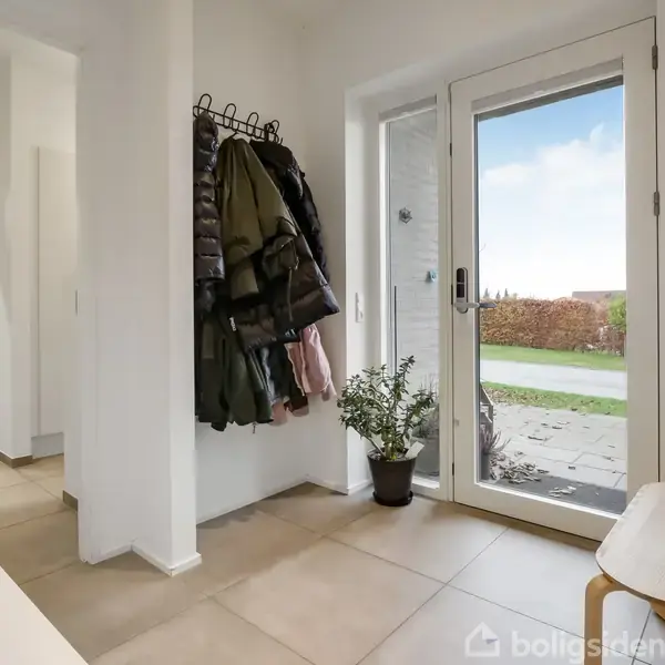 En entré med frakker hængende på en knagerække og en skammel ved siden af en glasdør. Udenfor ses en flisebelagt sti og grønne omgivelser.