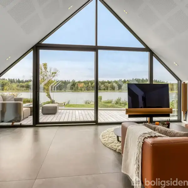 En stue med lædersofaer, fladskærms-tv og træskænk, vendt mod store glasvinduer. Udsigt til terrassen og sø med træer i baggrunden, under en høj trekantet loftform.