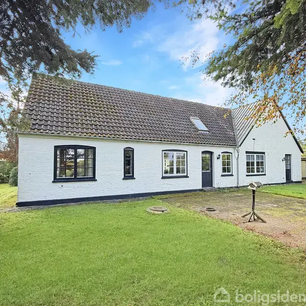 Et hvidt hus med mørke vinduesrammer står stille i en grøn have omgivet af træer. Huset har et mørkt skråt tag og en flisebelagt indkørsel.