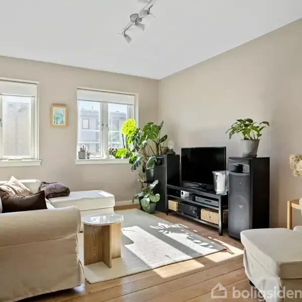 Stue med beige sofa og lænestole vender mod TV på møbelreol. Flere grønne planter pynter rummet. Lys strømmer ind fra to vinduer, der skaber en lys atmosfære.