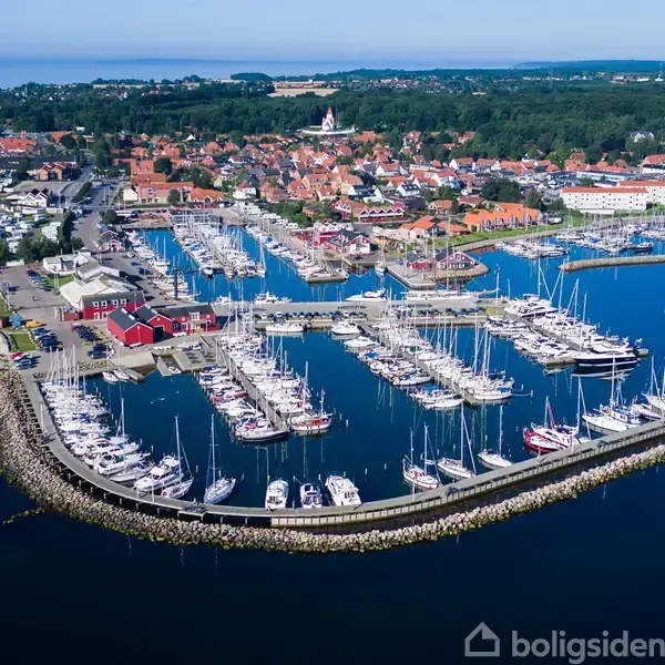 Havneanlæg fyldt med sejlbåde ligger ved kysten. Omgivet af en lille by med røde tage og grønne områder. Kystlinjen strækker sig mod horisonten.