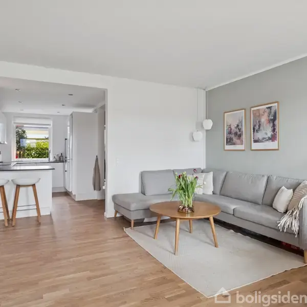 Grå sofa står i moderne stue, flankeret af træbord med blomster. Åbent køkken med barstole ses i baggrunden. Væggene har kunstbilleder, og rummet har lyst trægulv.