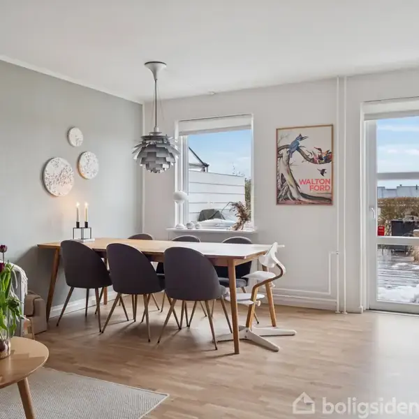 Et spisebord med seks stole står i en lys stue. En moderne lampe hænger over bordet. På væggen er der kunst og et skilt med teksten "WALTON FORD." Store vinduer viser en terrasse udenfor.