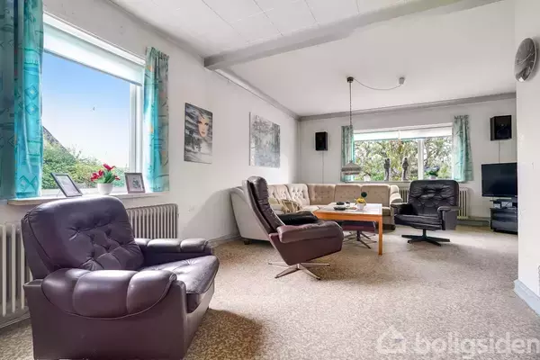 Stue med lænestole og sofa arrengeret omkring et sofabord. Væggene er dekoreret med plakater, og der er store vinduer med udsigt til træer. Lampe hænger fra loftet.