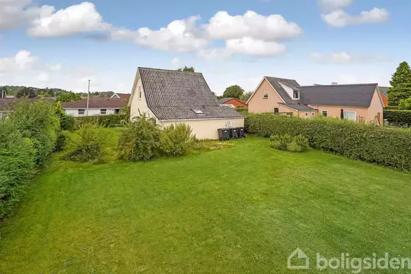 Et hus med skråtag står i en grøn have med hække. Flere huse ses i baggrunden under en blå himmel med enkelte skyer.