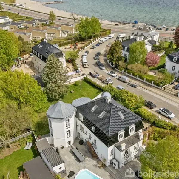 Hus med sort tag ligger i et grønt område nær en vej langs kysten. Flere biler kører på vejen. Der er udsigt over stranden og havet i baggrunden.