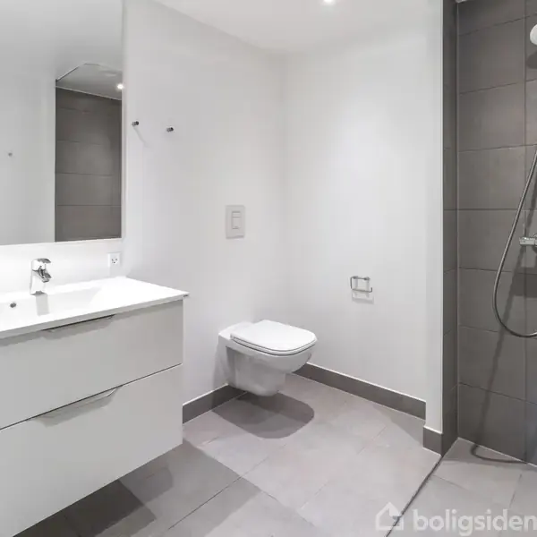 Hvidt badeværelse med væghængt toilet og bruser på en grå flisevæg. Et moderne vaskeskab med integreret spejl på en hvid væg.
