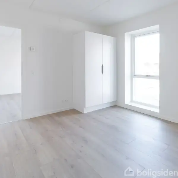 Et lyst, tomt værelse har en åben dør, en hvid garderobe, og et stort vindue. Rummet har lyse trægulve og hvide vægge, hvilket skaber en minimalistisk atmosfære.