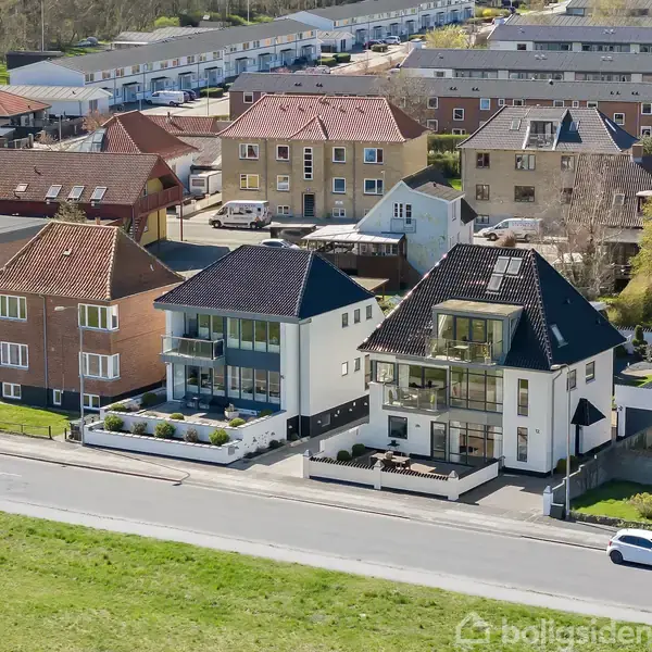 Tre moderne huse står i række langs en vej, omgivet af en række ældre boligblokke i en forstadsagtig bydel. To biler kører langs vejen foran husene.