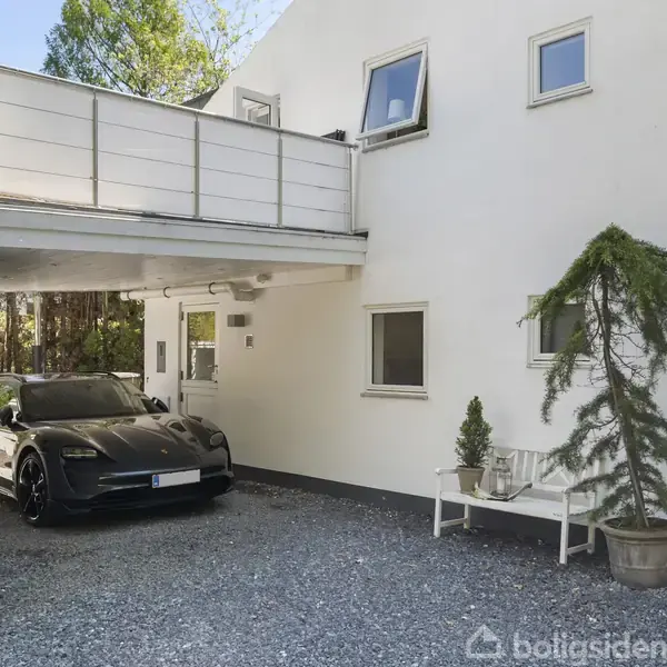 En sort bil holder parkeret under en carport ved siden af et hvidt moderne hus. Et lille træ står i en potte ved siden af en bænk. Tekst: "41B".