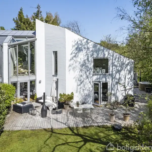 Hvidt, moderne hus med store vinduespartier, omgivet af frodig have med træer og buske. Terrassen har siddepladser og planter i krukker, der skaber en indbydende udendørs atmosfære.