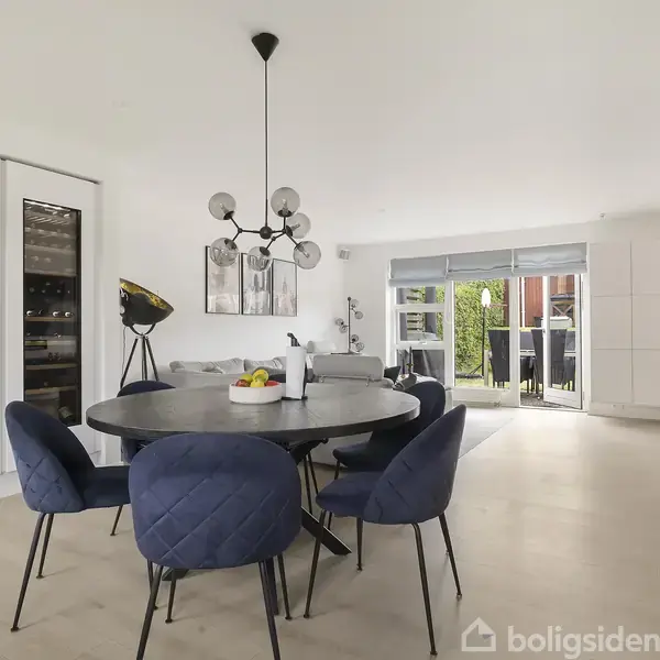 Rundt spisebord med blå, polstrede stole står i et moderne køkken-alrum. Der hænger en moderne lysekrone over bordet. Store vinduer åbner ud til en terrasse med grønne omgivelser.
