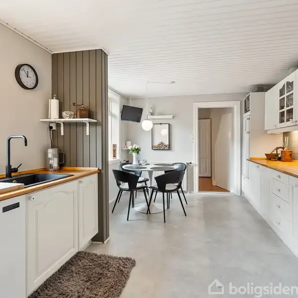 Køkken med hvide skabe og træbordplade indeholder opvaskemaskine, sort vandhane og vask, samt grå tæppe på gulvet. Rundt spisebord med sorte stole står i baggrunden under loftlampe.
