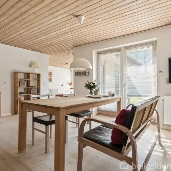 Gråt hjørnesofa sætter sig i stue med lyst trægulv, minimalistisk reol, og stort vindue. En træbordlampe står ved siden af en lav reol under et loft med træpaneler.