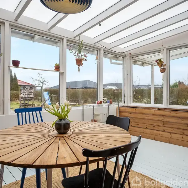 Rundt spisebord med stole står i en lys udestue. Bordet har en plante som dekoration. Omkring ses vinduer med potteplanter hængende, og udenfor er der en have.