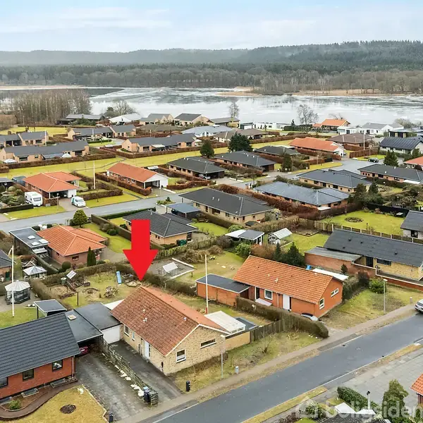 Rød pil peger på et murstenshus med rødt tag. Huset ligger i en villaby omkranset af flere huse og græsområder. En vej ses i forgrunden; skove ses i baggrunden.