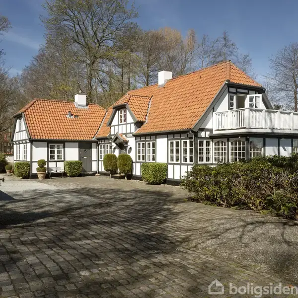 Hus med hvide vægge og sorte bjælker, rødt tegltag, beliggende i en skovomgivelse med brostensbelagt gårdsplads og buske. Det er forår med træer uden blade i baggrunden.