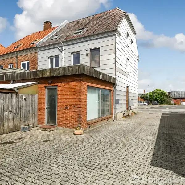 Et to-etagers hus med forskellige facadematerialer står i en gårdsplads. Omkringliggende bygninger og hegn omkranser pladsen, og himlen er skyet.