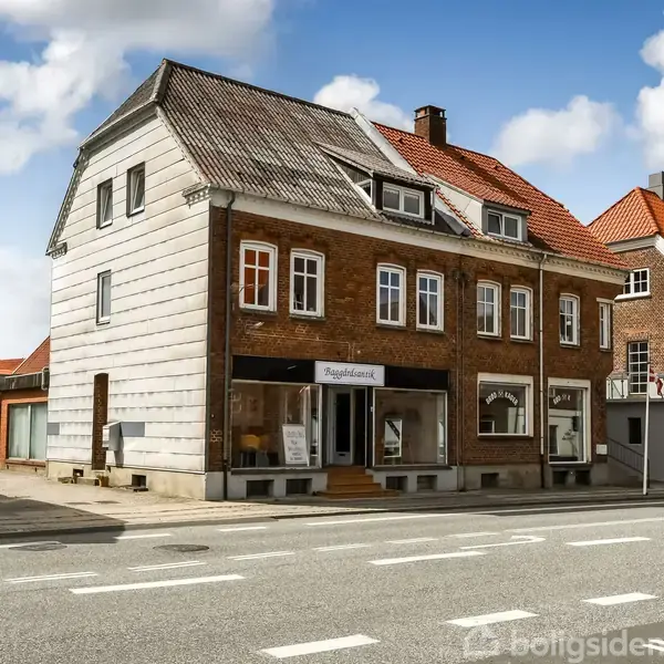 En to-etagers murstensbygning med butikker i stueetagen ligger langs en vej med fortov. Byens gadenavn på et skilt er "Nørregade." Skyer ses på den blå himmel.