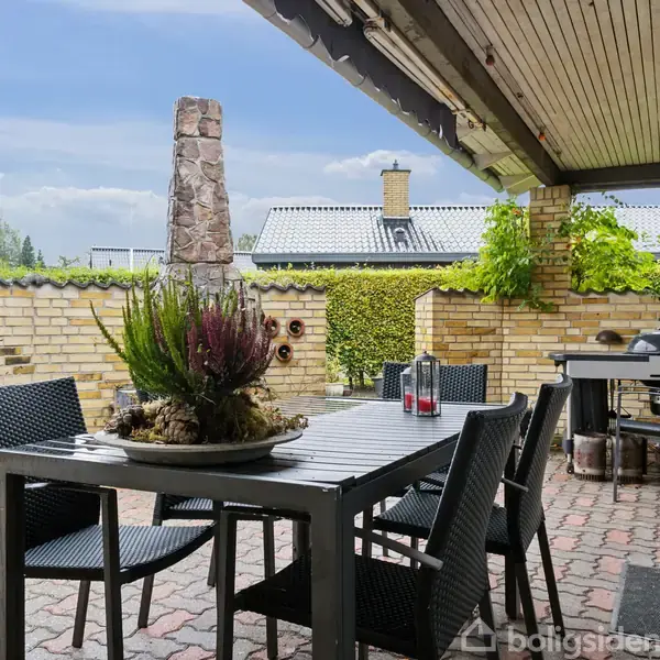 Terrasse med sort spisebord og seks stole actions. Potteplanter står på bordet. En grill står i hjørnet, omgivet af mursten og overdækket tag, med grønne klatreplanter på væggene.