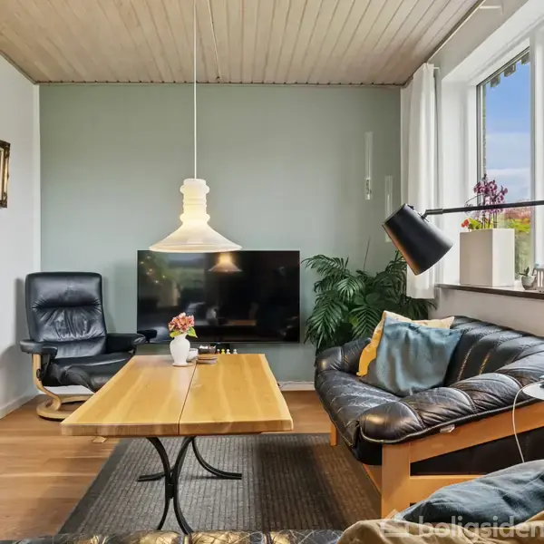 En stue med en sort lædersofa og lænestol, et træbord, fladskærm på væggen, grøn plante og store vinduer med udsigt til en terrasse.