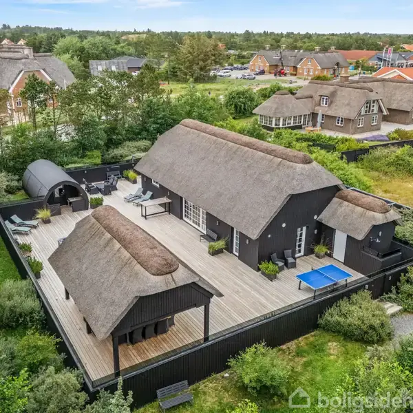 Sort stråtækt hus står på en træterrasse med udendørs møbler; beliggende i en landsby med flere lignende huse og grønne omgivelser.