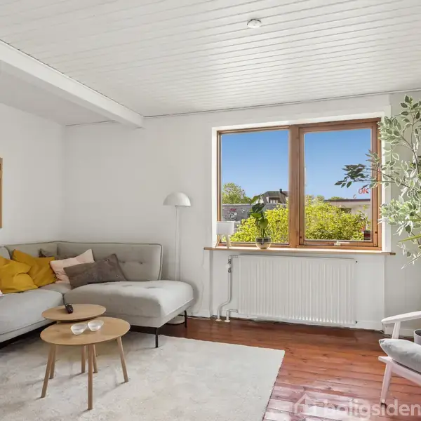 Sofa med puder står ved et vindue, der viser blå himmel og grønne træer. Et lille sofabord og en gulvlampe er ved siden af. Vægplakater fuldender det lyse rum.