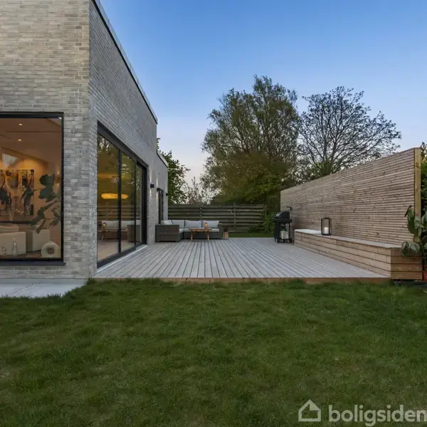 Et moderne hus med store vinduer, der afslører en stue indeni, står ved siden af en træterrasse med havemøbler og grill i en frodig have med træer i baggrunden.