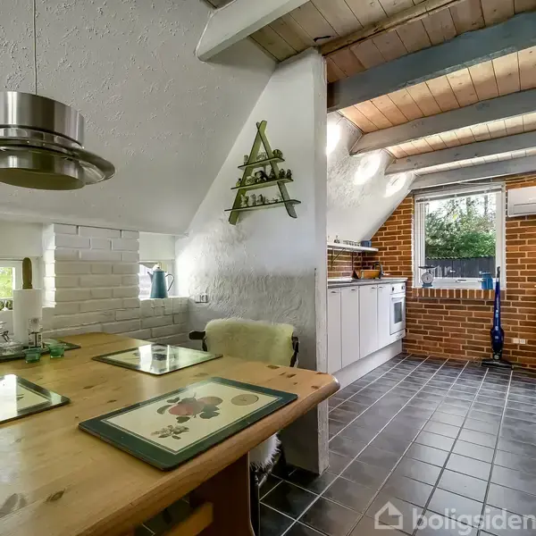 Et køkkenbord prydet med blomsterunderlag står ved siden af en hvid murstensvæg, mens et sort flisegulv strækker sig ind i rummet med brune murstensvægge og trælofter.