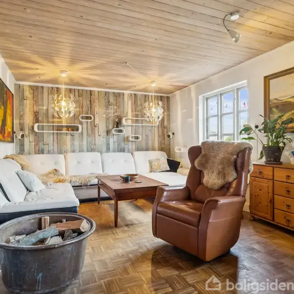 En komfortabel stue med en hvid sofa og lænestol står ved et sofabord. Væggene har farverige malerier, og der er en kamin til træbrænde. En kommode med planter står under et vindue.