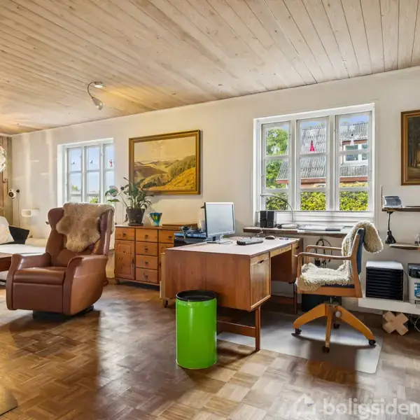 Et hyggeligt dagligstuekontor med en lænestol og et træskrivebord fyldt med papirer og kontorgenstande. To malerier hænger på væggen over et parketgulv og grønne planter.