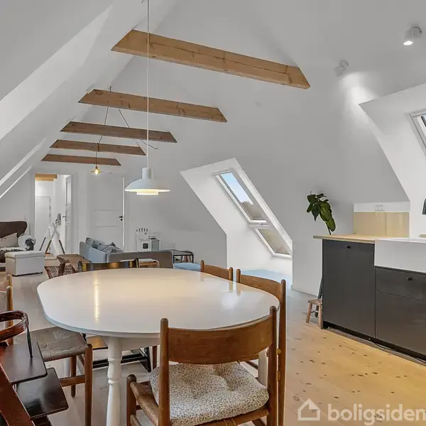 Et alrum med skrå vægge og synlige træloftsbjælker indeholder et spisebord med stole. I baggrunden er et køkken med ovenlysvindue. Et enkelt væghængt grønt plante dekorerer rummet.