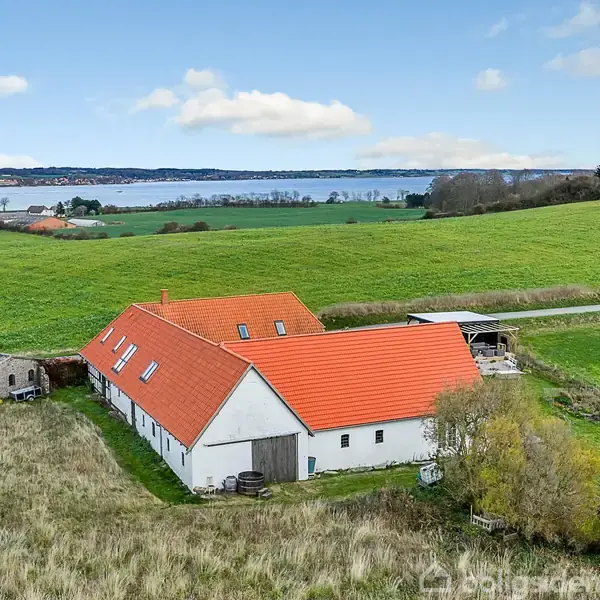 En hvid bygning med rødt tag ligger i et grønt landskab nær en sø. Omgivet af marker og bakker, ses også en mindre stenbygning.