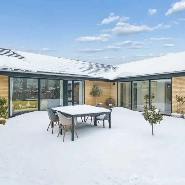 Terrassebord omringet af stole står stille, dækket af let sne i en gårdhave. Bygningen, gul mursten med store vinduer, er omkranset af sneklædte tage.