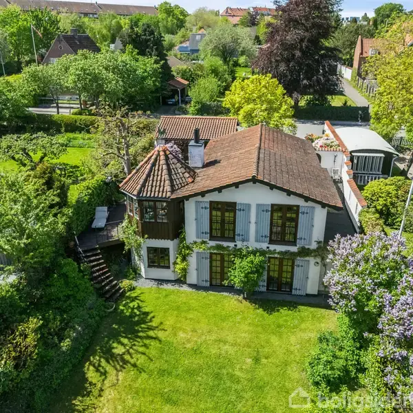 Et hus med rødt tegltag omgivet af grønne træer. Der er en terrasse med møbler ved siden af huset. Omgivelserne inkluderer flere huse og en frodig have.