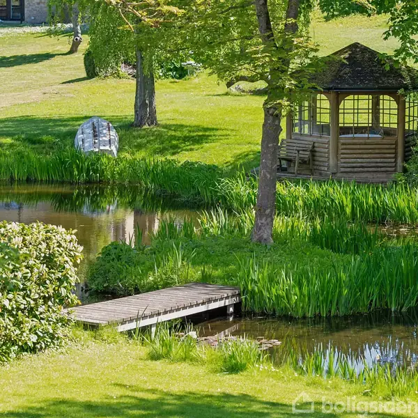 En lyseblå sofa står i en karnap foran store vinduer, omgivet af grøn udsigt. En rund hvid sofabord med blomster og magasiner står foran sofaen på et lyst tæppe.