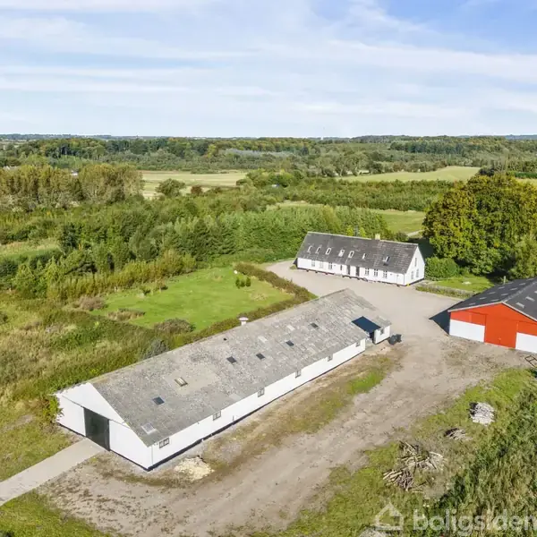 Længere bygning står som hovedfokus, omgivet af åben mark og træer. En mindre rød bygning står ved siden af. Landskabet er grønt og frodigt under en blå himmel.